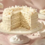white chocolate coconut cake recipe photo wcccr with snowy white frosting and coconut coating on an ivory cake stand, showcasing a removed slice revealing moist layers