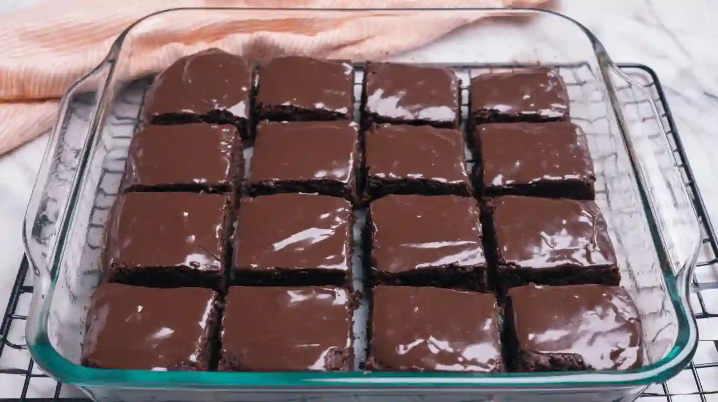 protein-brownies-serving2 A glass baking dish filled with neatly cut squares of glossy chocolate protein brownies, placed on a cooling rack with a striped towel in the background.