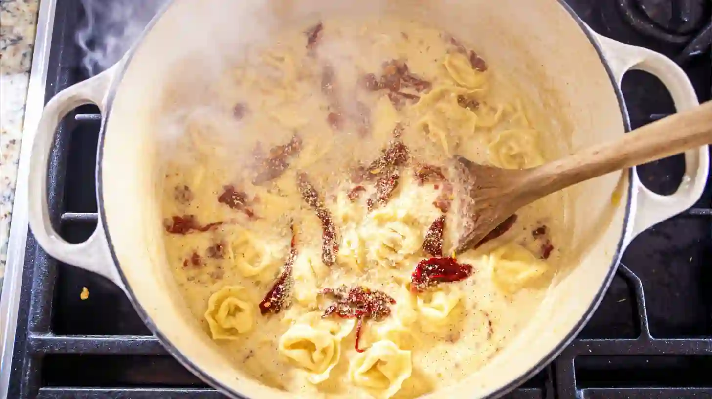 creamy-garlic-tortellini-cream-sauce-step5 stirring heavy cream, parmesan, and sun-dried tomatoes into tortellini for creamy garlic shrimp pasta