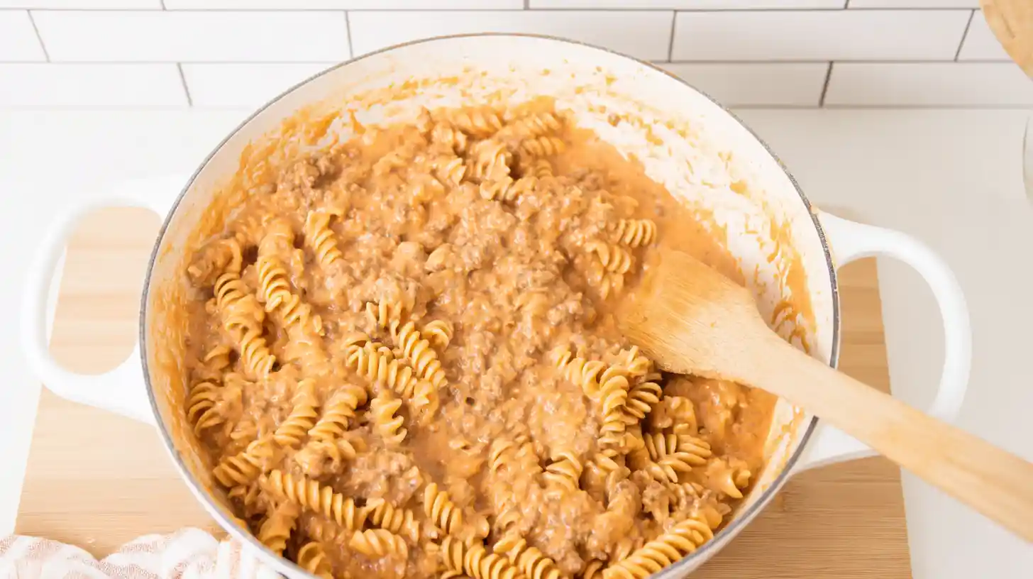 step5-fold-rotini-noodles-cheesy-beef-casserole-recipe Folding rotini noodles into creamy beef and cheddar sauce – one-pot dinner idea for busy families