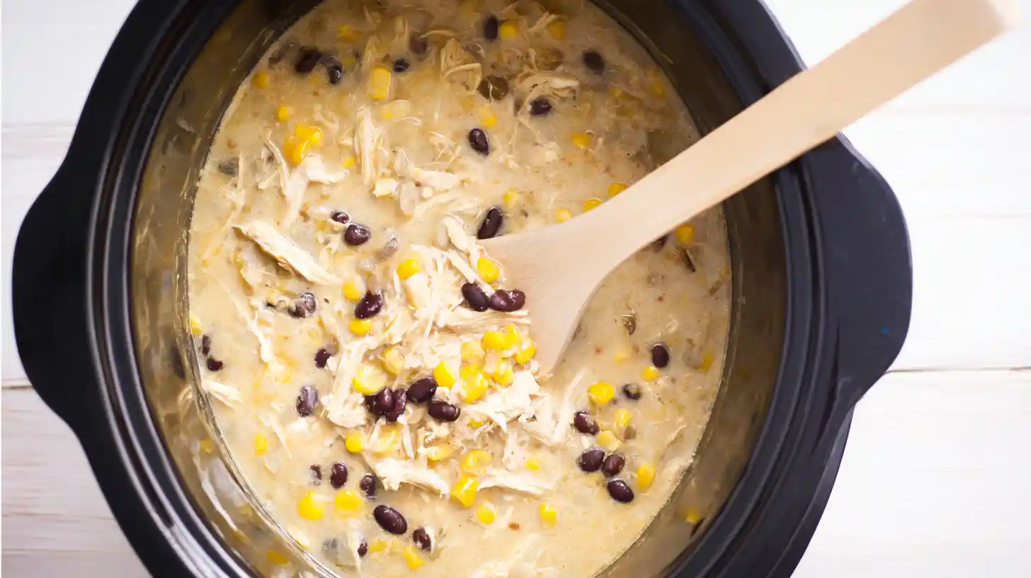 Stirring black beans and corn into creamy green enchilada chicken soup in a Crock Pot – hearty slow cooker chicken soup with protein and fiber
