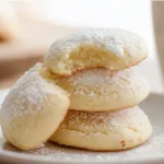 Soft homemade cream cheese cookies stacked on a white plate, lightly dusted with powdered sugar, with a bite revealing their fluffy, buttery interior.