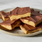 Stacked 6 Ingredients easy No-Bake Chocolate Peanut Butter Bars with a rich chocolate topping and creamy peanut butter base on a white plate, photographed on a white marble background in natural lighting. Easy homemade dessert recipe image.